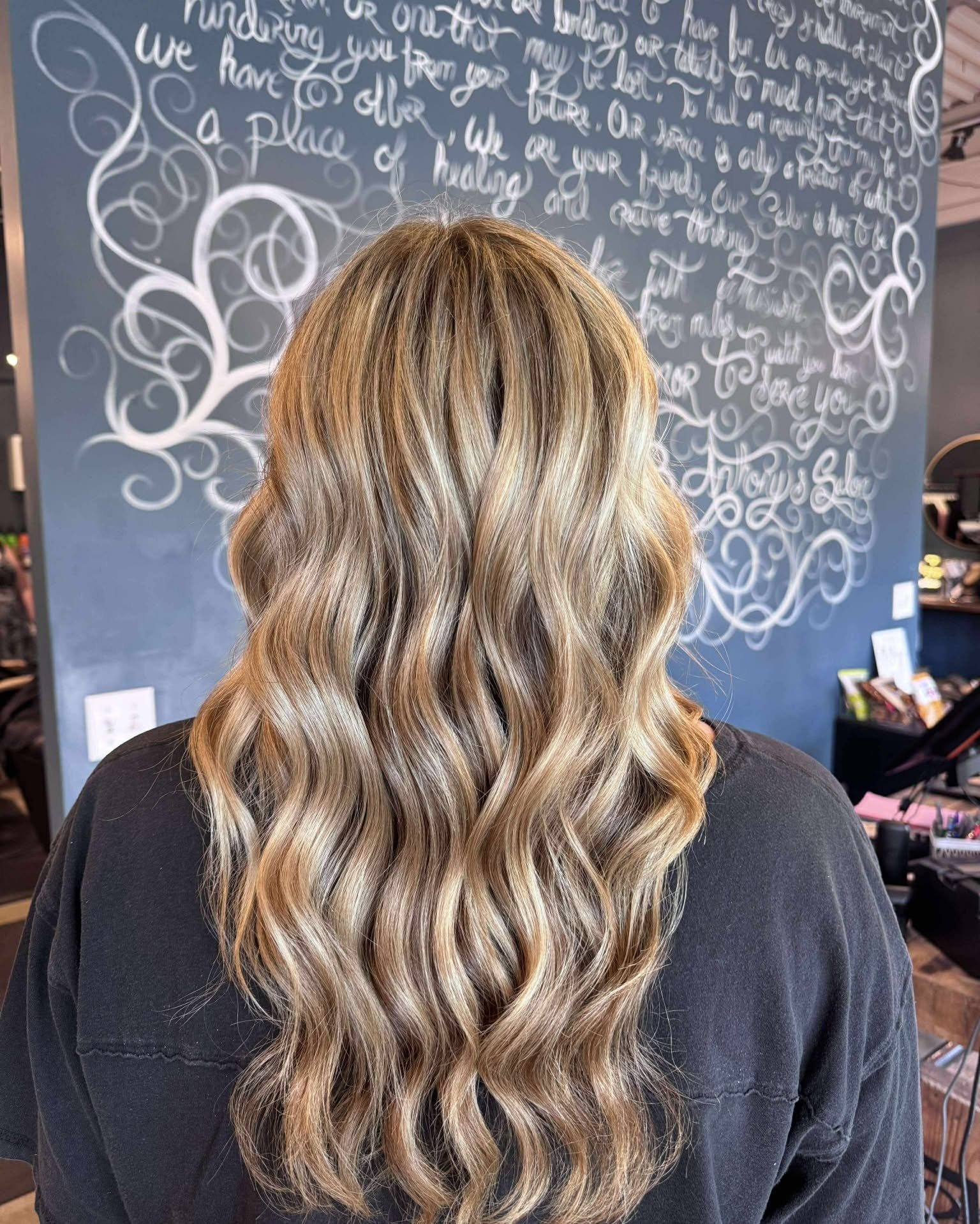 Long wavy blonde balayage against the salon's hand-lettered feature wall